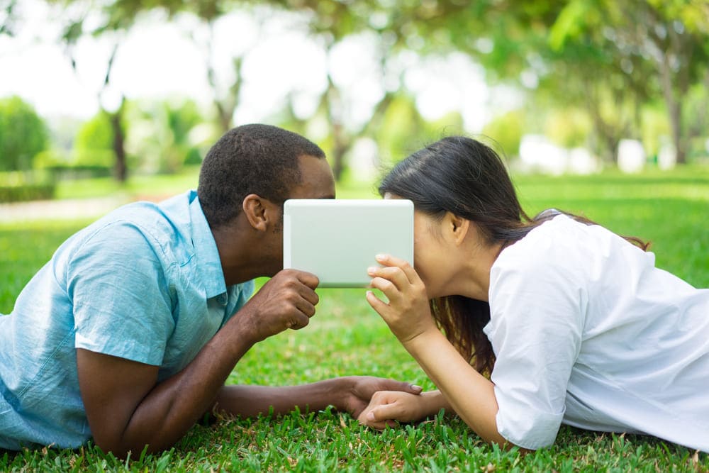 heureux couple multiethnique couche sur l herbe se cachant les visages derriere le touchpad