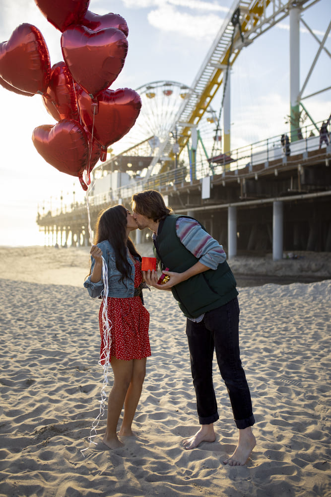 jeune couple sur la plage au parc d attractions tenant des ballons en forme de coeur et une boite cadeau tout en s embrassant1